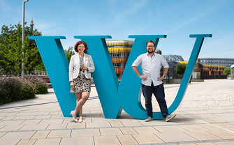 Univ.Prof. Sigrid Stagl vom Department für Sozioökonomie und Jonathan Krupitza von der Personalabteilung lehnen an einem großen WU-Logo.