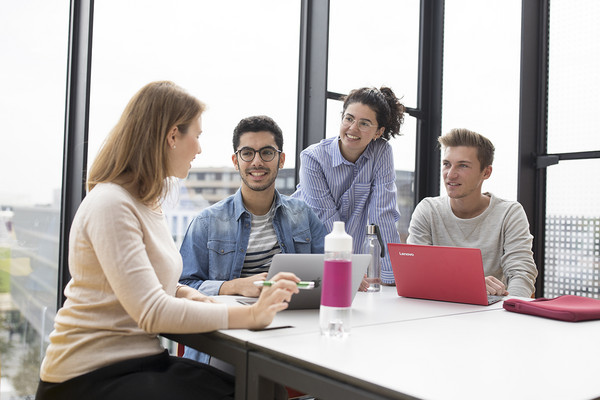 2 students sit at a table with their laptops and talk to each other