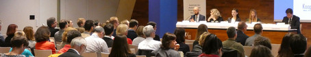 Zuhörer bei einem Symposium. Dahinter Referenten hinter einem Podium sitzend.