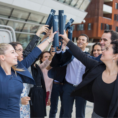 A group of students stand together in front of the library building and hold their diplomas up to the air