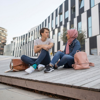 Ein Student und eine Studentin sitzen auf einer Holzbank vor einem Universitätsgebäude und unterhalten sich