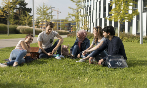 Fünf Studierende sitzen im Halbkreis in der Wiese vor dem D2