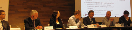 People are sitting at a long table, listening to a man seated in the middle. In front of them are name signs and microphones.