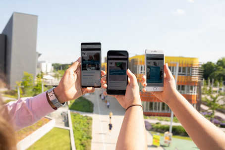 Drei Personen halten ihre Mobiltelefone gemeinsam in die Höhe, auf den Telefonen ist die WU Website geöffnet, im Hintergrund der WU Campus.