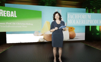Christina Holweg, a standing woman with moderator cards in her hand in a recording studio. The background shows the words: Regal and Fachforum Molkereiprodukte. 