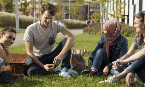 Fünf Studierende sitzen im Halbkreis in der Wiese vor dem D2