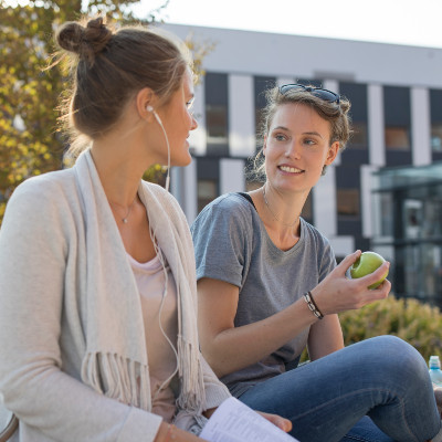 Zwei Studentinnen sitzen auf einer Holzbank vor dem Gebäude D2 und unterhalten sich.