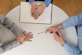 Drei Personen an einem Tisch. Erkennbar sind nur die Hände. Eine Hand weist mit einem Kugelschreibe auf das Gegenüber.