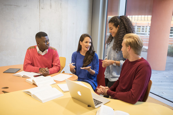 Students sit at a table. On the table there are study materials and an opened laptop 