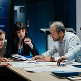 [Translate to English:] Vier Menschen sitzen stehen im Büro um einen Tisch