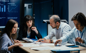 [Translate to English:] Vier Menschen sitzen stehen im Büro um einen Tisch