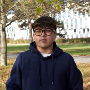 Portrait von Ali Akbar Abdramanov vor einem herbstlichen Hintergrund im Prater. Er ist ein junger Mann mit kurzen, schwarzen Haaren und einer Brille in einem dunkelblauen WU-Hoodie.