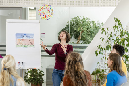 Studierende sitzen im Sesselkreis, Vortragende wirft einen Ball in die Luft und lächelt